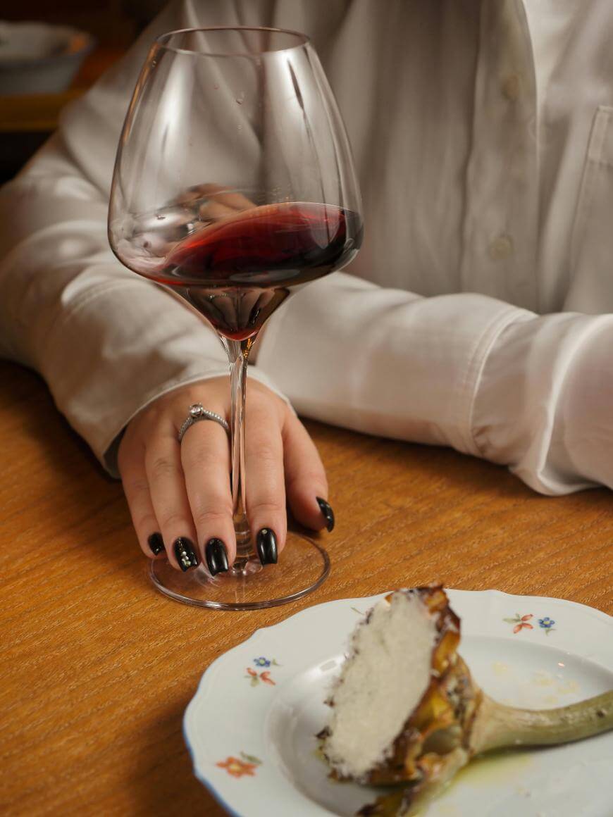 A person in a white shirt holds a glass of red wine on a wooden table, with a plate containing a partially eaten artichoke in the foreground. The person wears a ring and has black painted nails.