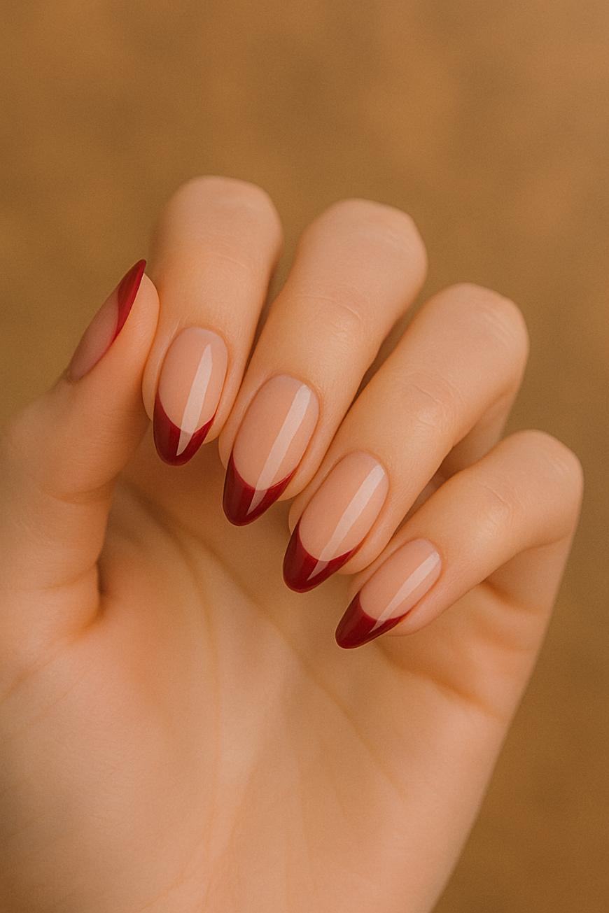 A hand with long, almond-shaped nails featuring a nude base and deep red, pointed French tips, displayed against a soft, brownish background.