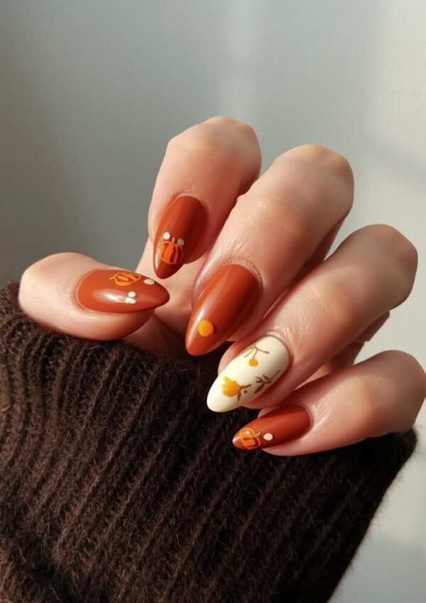 A hand with almond-shaped nails painted in autumn colors, featuring designs like pumpkins and leaves on a few nails. The person is wearing a brown knit sweater.