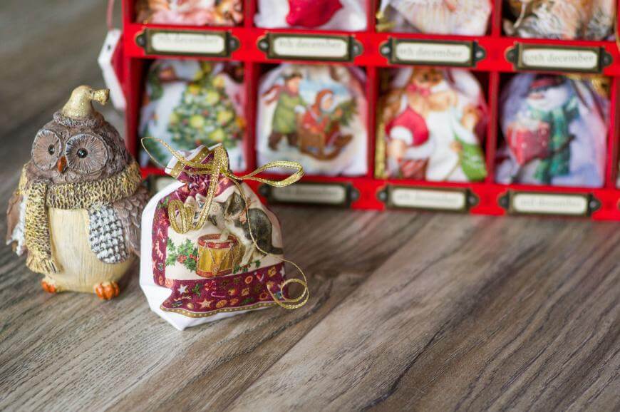 A decorative owl figurine stands next to a small gift bag with festive designs, in front of a red advent calendar filled with illustrated holiday pouches on a wooden surface.