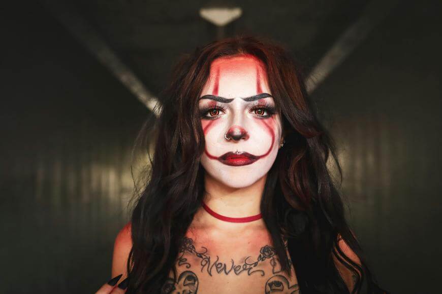 A woman with long dark hair and dramatic clown makeup, featuring red and black details, stands in a dimly lit hallway. She wears a choker and has visible tattoos on her chest.