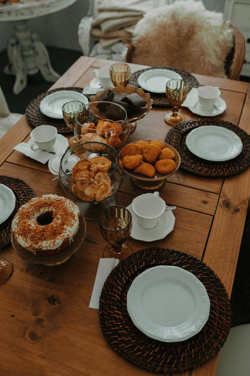 A wooden table set for four with white plates, cups, brown woven placemats, gold glasses, and a variety of baked goods including a bundt cake, pastries, muffins, and cookies in glass bowls.