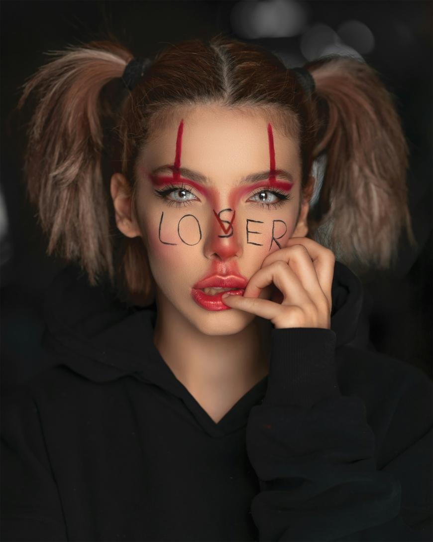 A person with pigtails and dramatic clown-like makeup poses, with LOSER written across their face in black letters. They wear a black hoodie and touch their lip, gazing intensely at the camera.