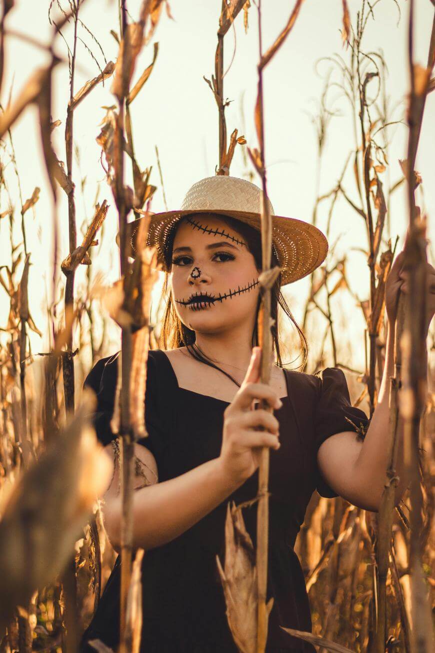 A person wearing a straw hat and black dress stands in a cornfield, with makeup resembling stitched scars on their face, creating a spooky, scarecrow-like appearance. Warm sunlight filters through the dry corn stalks.