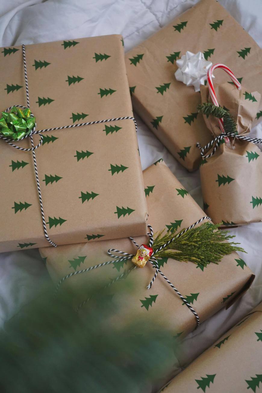 Five presents wrapped in brown paper decorated with small green Christmas trees, tied with black-and-white string, and adorned with festive accents like greenery, bows, a candy cane, and a holiday bell.