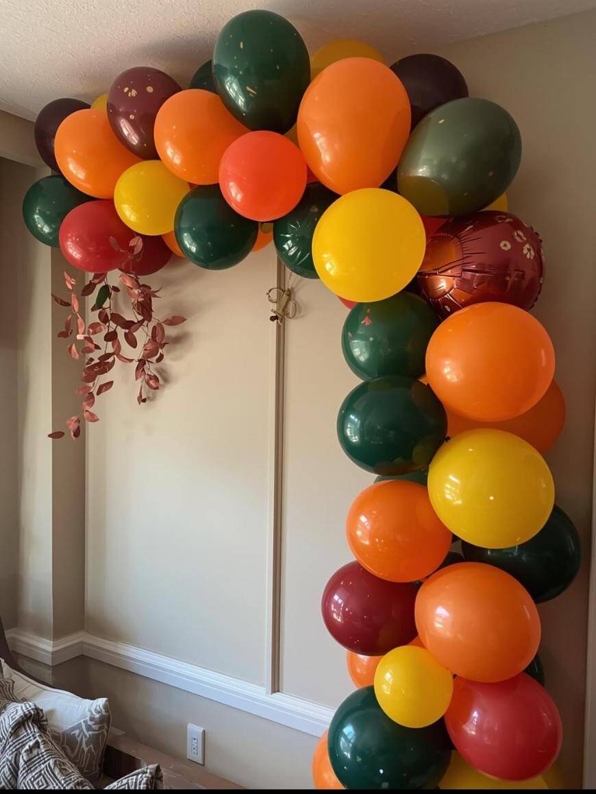 A colorful balloon arch decorates a corner of a room, featuring balloons in shades of orange, yellow, green, and maroon, with a small bunch of autumn leaves for accent.