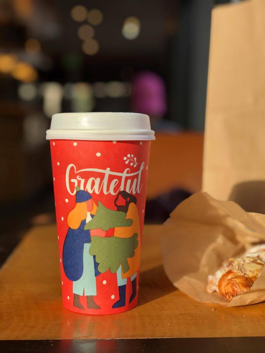 A red coffee cup with white dots and the word Grateful shows an illustration of three people hugging a green tree. The cup sits on a table next to a partially unwrapped pastry and a brown paper bag.