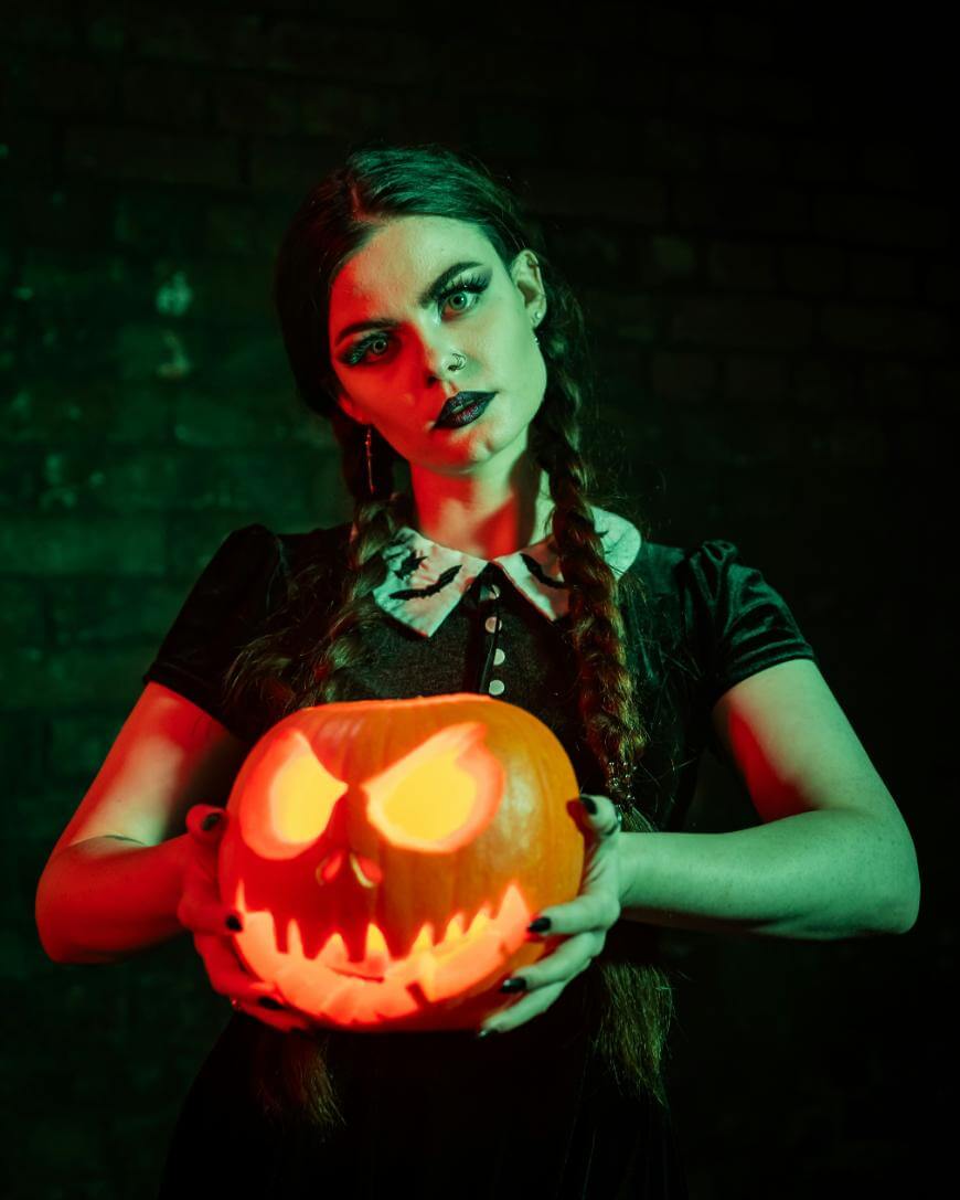 A woman with braided hair and dark makeup, dressed in a black dress with a white collar, holds a glowing, menacing jack-o’-lantern in front of her against a dark, moody background.