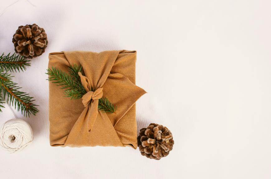 A gift wrapped in brown fabric with a pine branch, placed on a white surface next to pinecones, a ball of white string, and green fir branches.