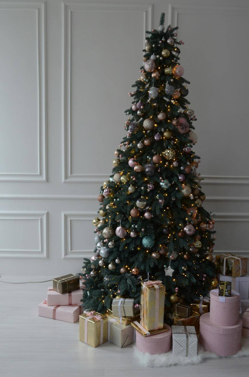 A decorated Christmas tree with gold and pink ornaments stands in a white room. Wrapped gifts in pink, gold, and white paper are arranged at the base of the tree.