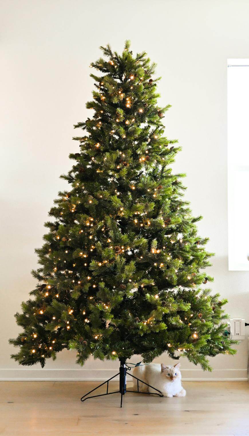A tall artificial Christmas tree with white lights stands in a bright room; a fluffy white cat is lying at the base of the tree near the stand.