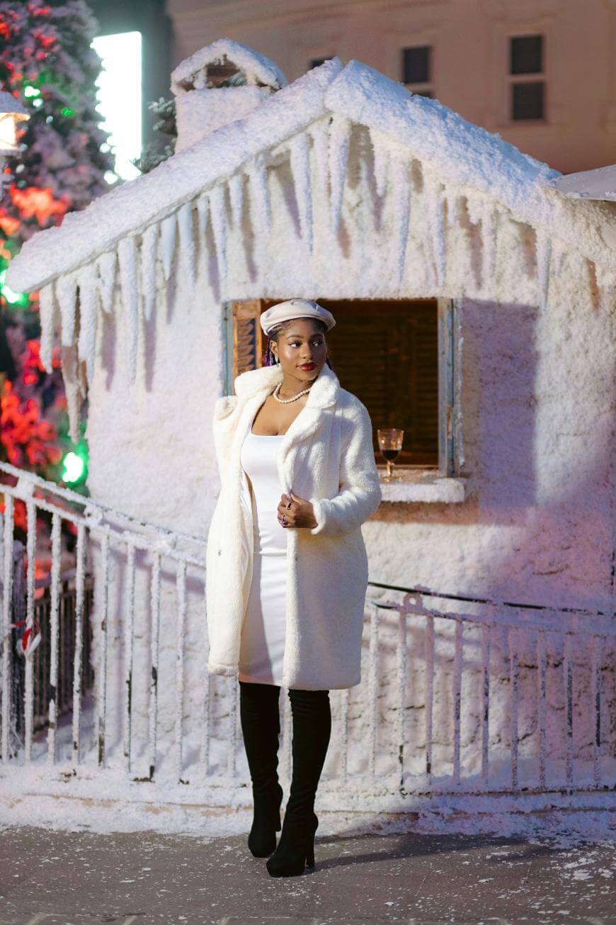 A woman in a white coat and dress stands in front of a small snow-covered building with icicles, wearing a white beret and black boots. The scene appears festive and wintry, with bright lights in the background.