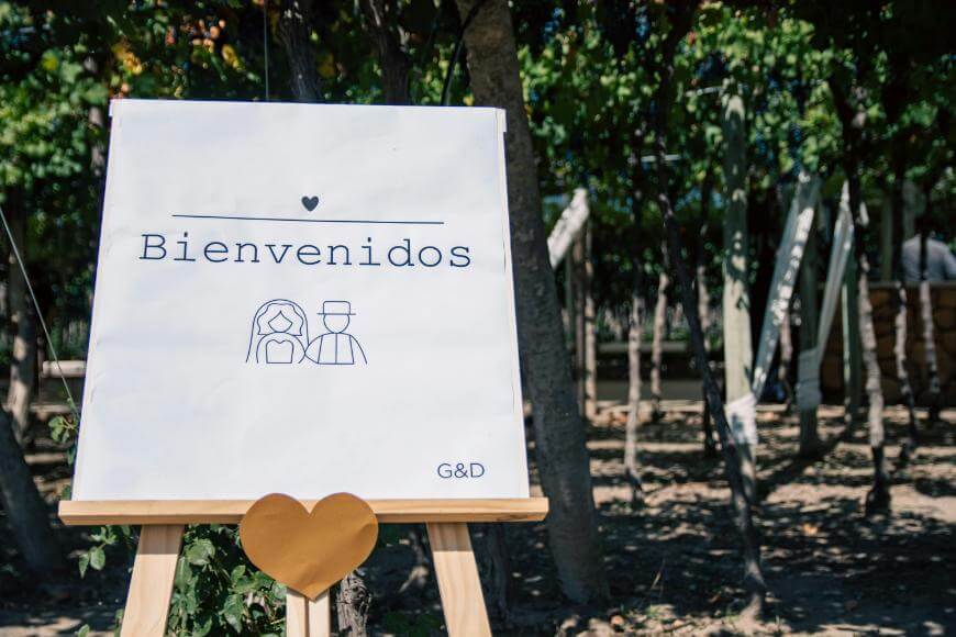 A white welcome sign with Bienvenidos and a simple drawing of a bride and groom stands outdoors among trees, decorated with a small heart and the initials G&D.