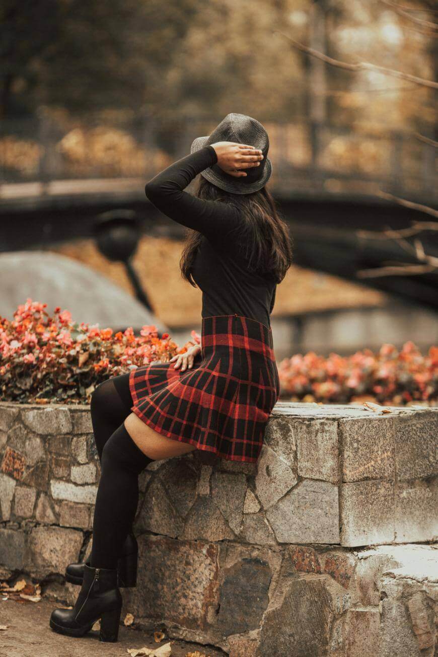 A woman in a black top, red plaid skirt, thigh-high socks, and boots sits on a stone wall, holding a hat on her head. She faces away from the camera, with autumn trees and a bridge in the background.