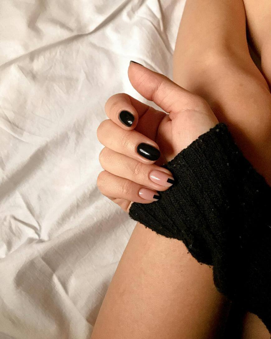 A hand with manicured nails, featuring black nail polish on some fingers and nude polish with black tips on others, rests on a bare leg. The person is wearing a black long-sleeved top and is sitting on white bedding.