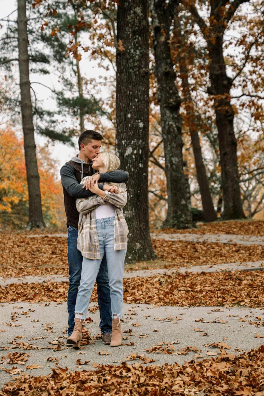 A couple stands in an autumn park, surrounded by fallen leaves. The man hugs and kisses the woman on the forehead while she smiles and holds his arms, both dressed in casual fall clothing. Tall trees with orange leaves are behind them.