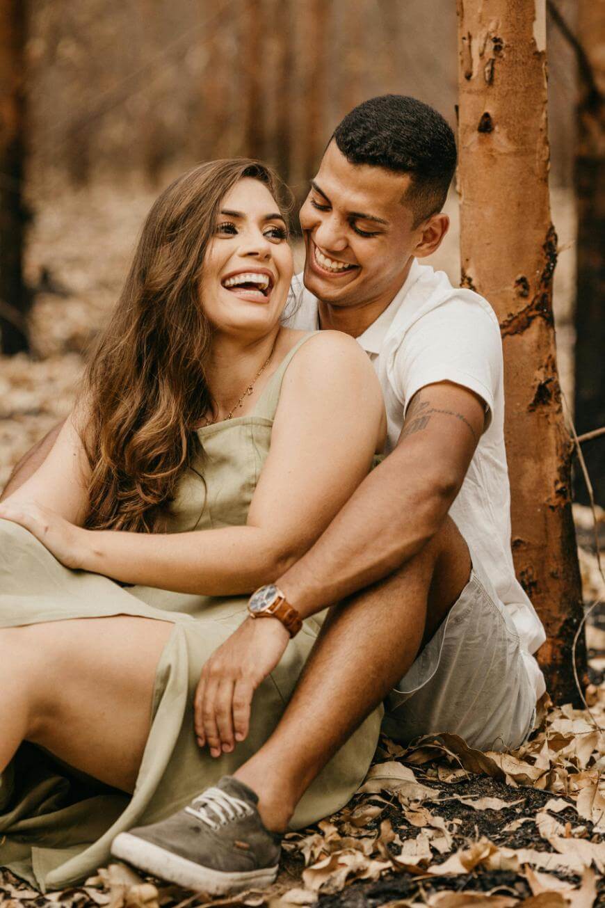 A smiling couple sits on the ground in a forest with brown tree trunks and dry leaves. The woman wears a green dress, and the man, in a white shirt, sits behind her, hugging her and both look happy and relaxed.