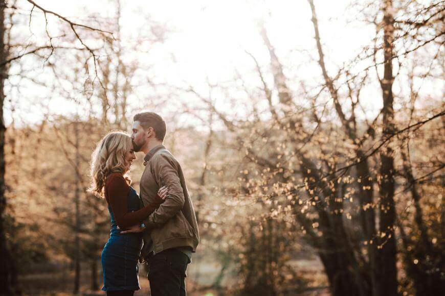 A man kisses a womans forehead as they embrace in a sunlit forest. They stand close together, surrounded by leafless trees and golden light filtering through the branches.