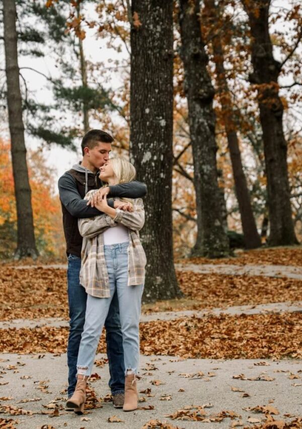 A couple stands in an autumn park covered in fallen leaves. The man hugs and kisses the woman on the forehead while she smiles and holds his arms. Both are dressed in casual fall clothing and surrounded by tall trees with orange foliage.