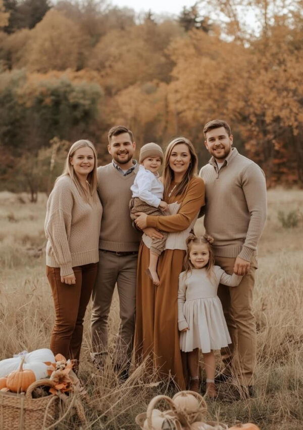 A family of six, including two young children, stands together in a grassy field with autumn trees in the background. They wear coordinated neutral outfits and smile at the camera. Pumpkins and baskets sit in the foreground.