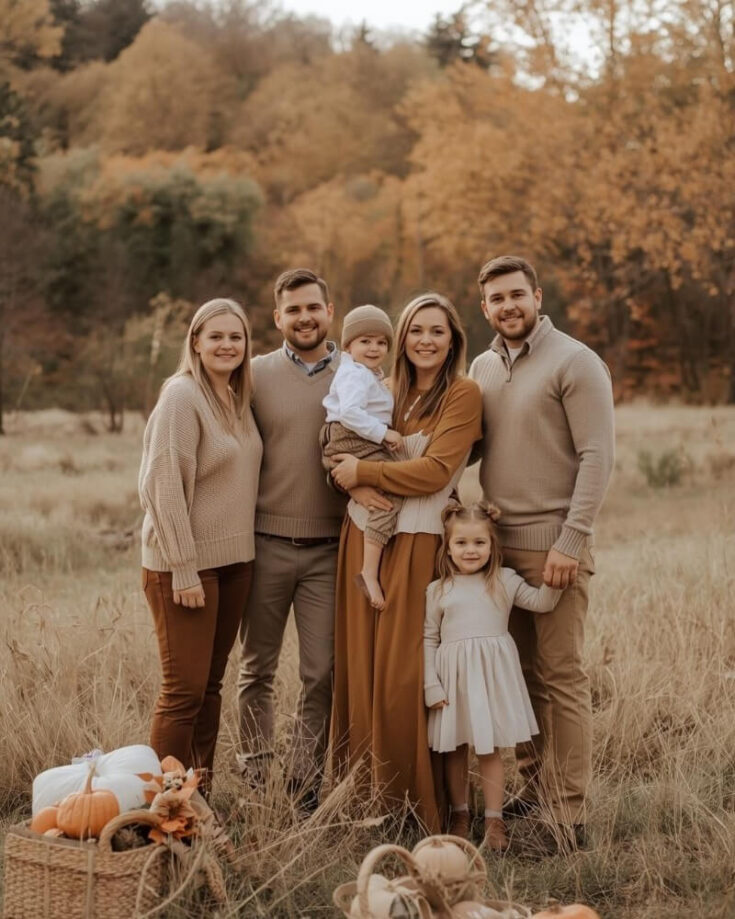 A family of six, including two young children, stands together in a grassy field with autumn trees in the background. They wear coordinated neutral outfits and smile at the camera. Pumpkins and baskets sit in the foreground.