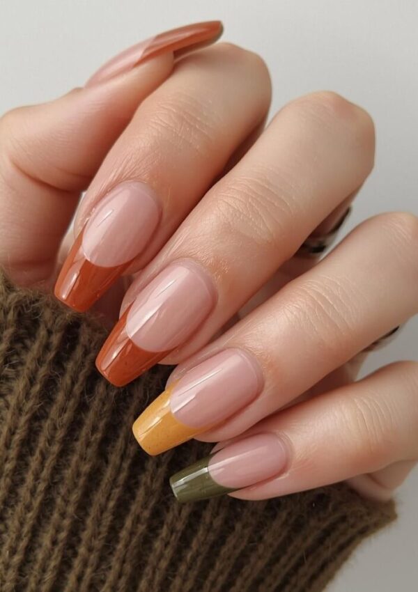 A hand with long, square-shaped nails featuring a French manicure in fall colors: orange, amber, mustard yellow, and olive green tips, against a neutral background. The person is wearing a brown knit sweater.