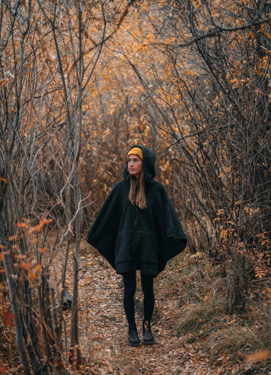 A woman wearing a dark poncho, black tights, boots, and a yellow hat stands on a leaf-covered path surrounded by autumn trees with orange and brown leaves.