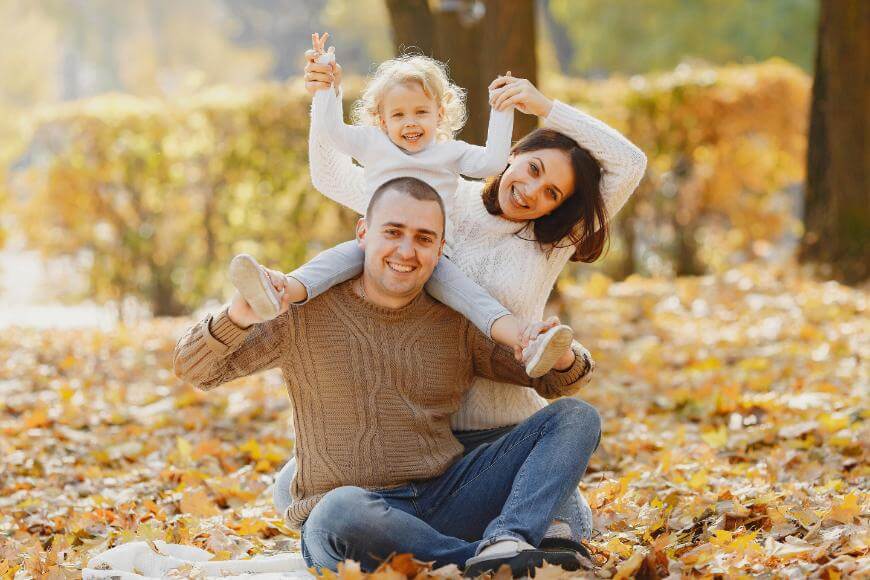 A smiling family sits on autumn leaves in a park. A man sits cross-legged, a young child sits on his shoulders holding hands with a woman who is kneeling beside them. All are wearing cozy sweaters and look happy.