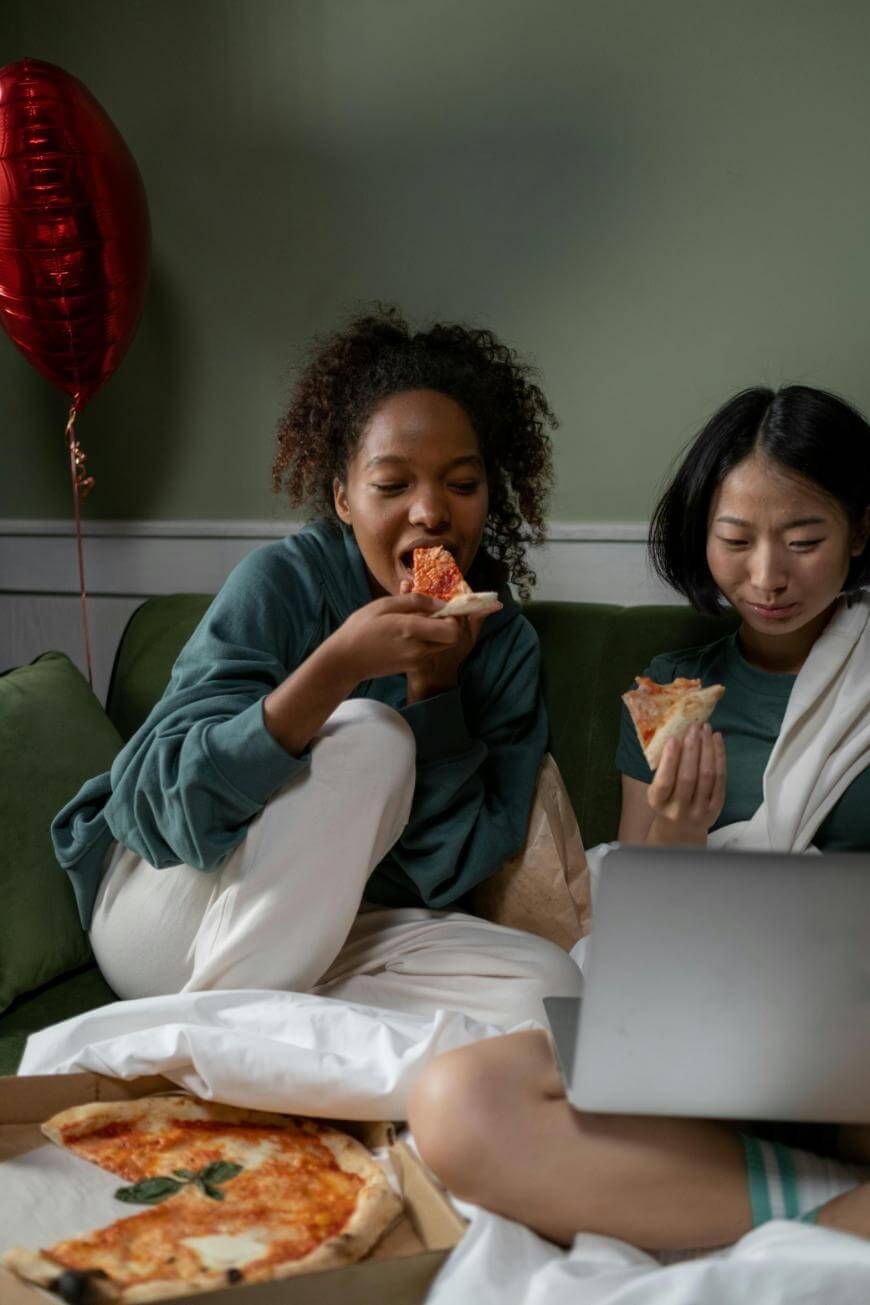 Two women sit together on a couch, eating pizza and watching something on a laptop, with a red heart-shaped balloon in the background and a pizza box open in front of them.