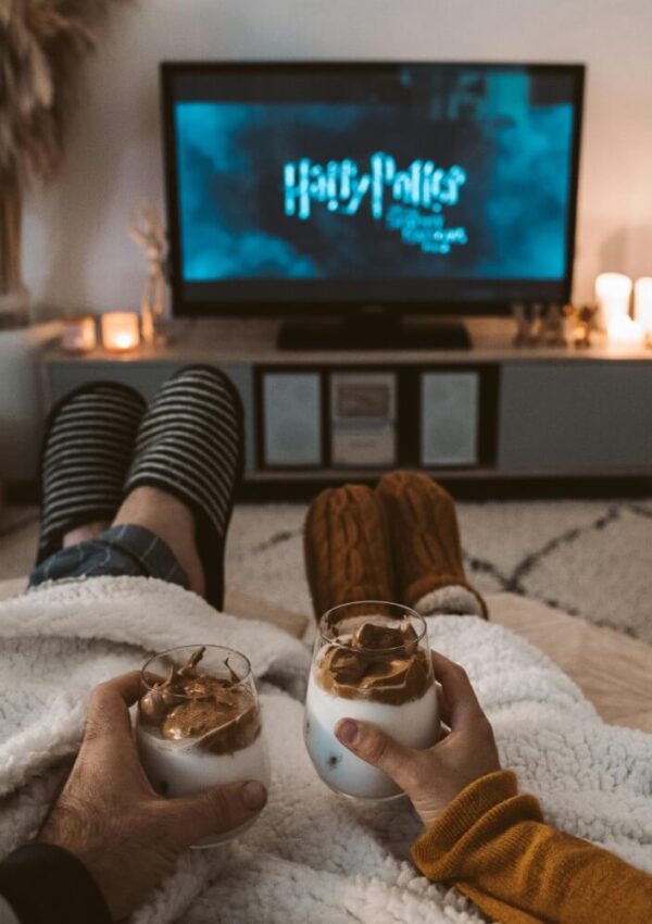 Two people with cozy slippers relax under a blanket, holding whipped coffee drinks, while watching a Harry Potter movie on TV in a softly lit living room.