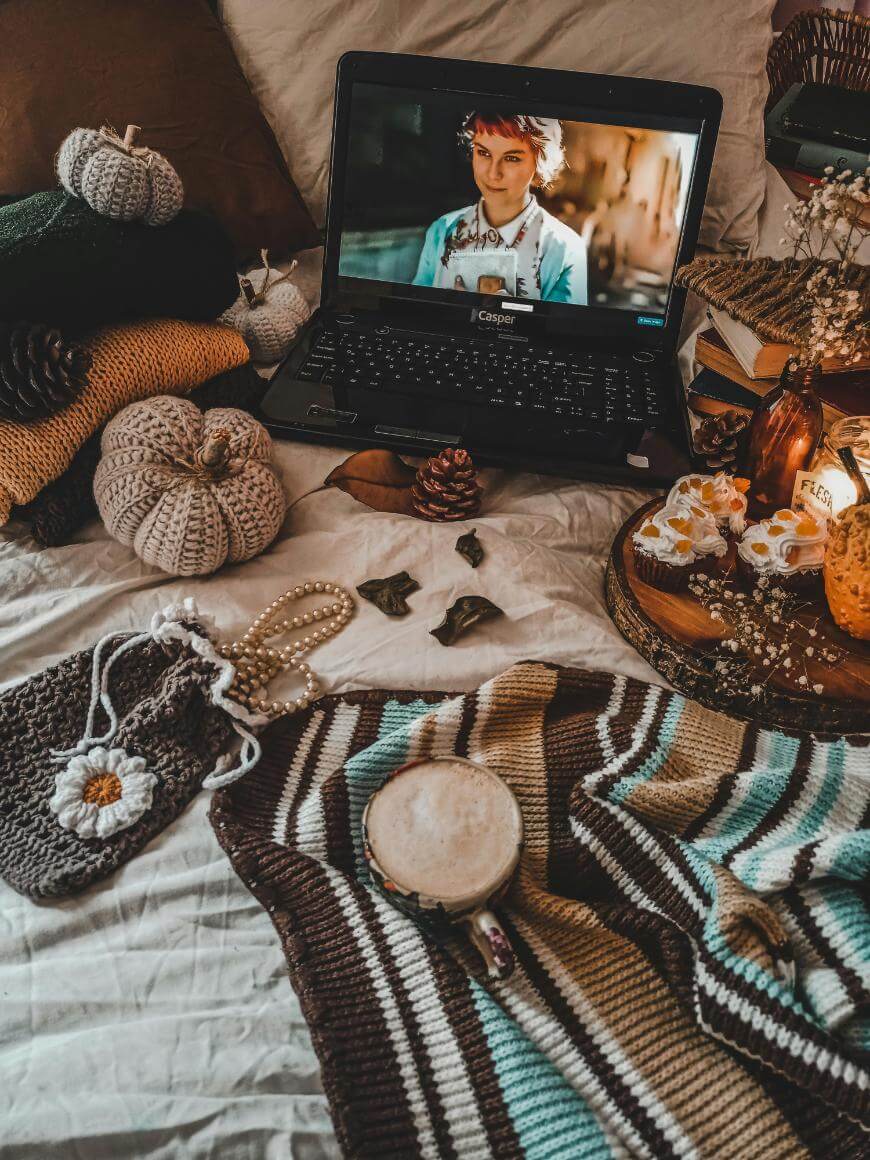 A cozy scene with a laptop playing a movie on a bed, surrounded by knitted pumpkins, fall decor, a coffee cup, a crocheted pouch, a pearl necklace, and a striped blanket.