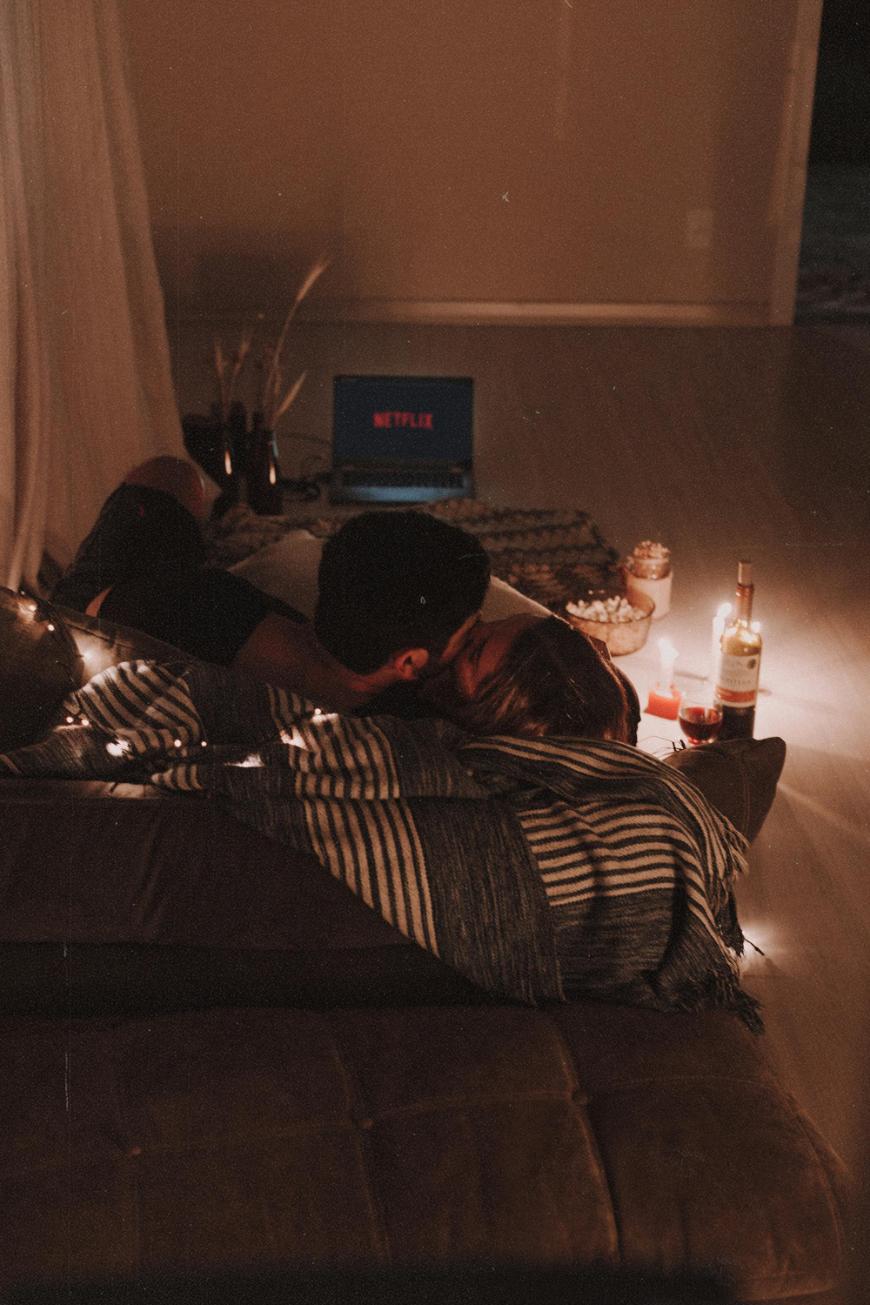 A couple cuddles on a couch under a striped blanket, surrounded by dim string lights, snacks, wine, and glasses. A laptop with the Netflix logo is open in the background, creating a cozy movie night atmosphere.