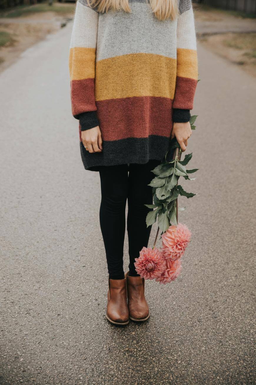 A person wearing a striped sweater, black leggings, and brown boots stands on a paved road, holding pink flowers upside down by their side. The persons face is not visible.