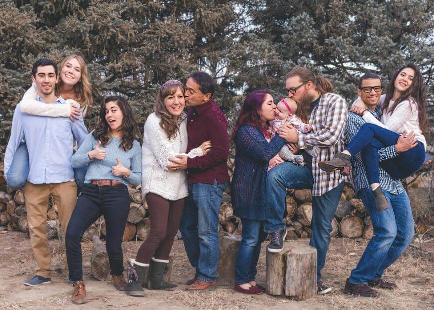 A group of nine people pose happily outdoors in front of trees and logs. Some couples embrace, one person holds a baby, and everyone is smiling or laughing, wearing casual fall or winter clothes.