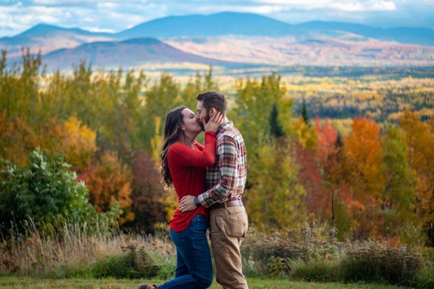 A couple stands kissing in front of vibrant autumn foliage, with colorful trees and rolling mountains in the background under a partly cloudy sky.