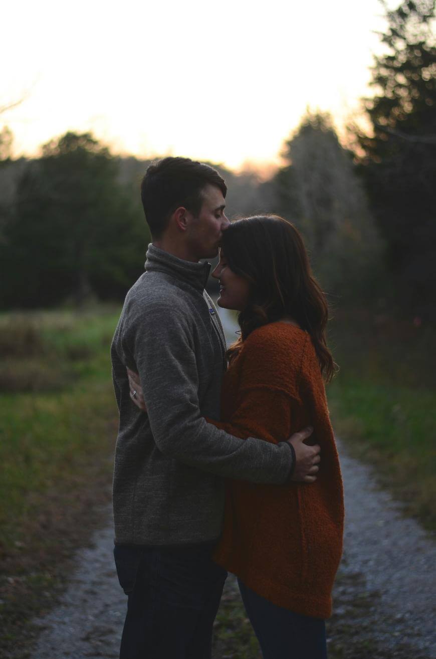 A man and woman stand close on a gravel path, embracing as the man kisses the woman’s forehead. They are outdoors with trees and soft evening light in the background.