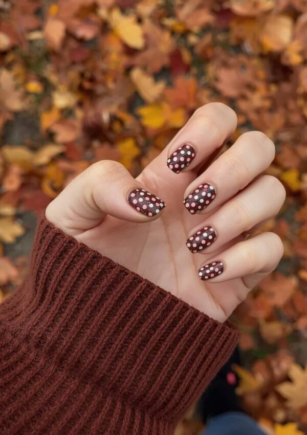 A hand with brown nail polish and white polka dots holds up against a background of autumn leaves. The person is wearing a brown knit sweater.