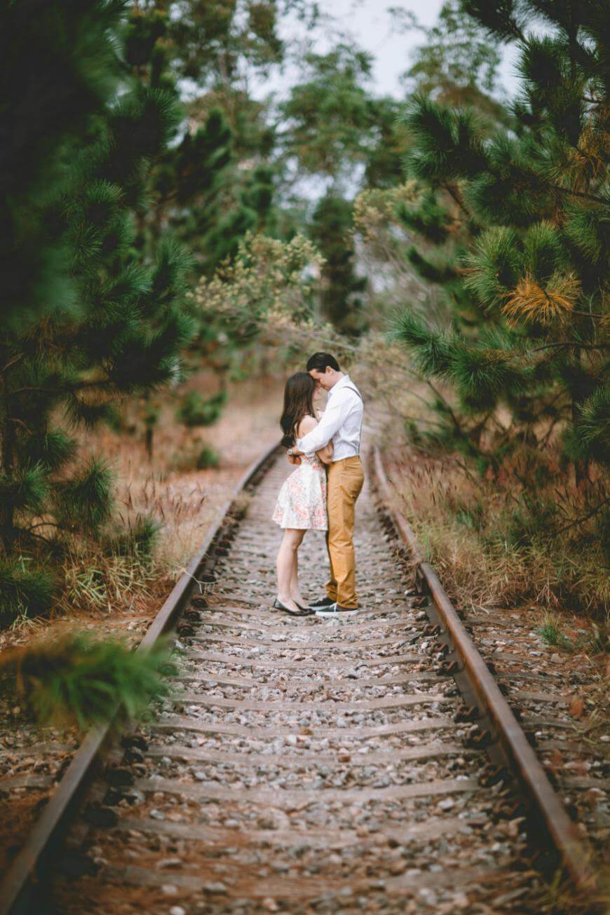 A couple embraces while standing face to face on a railway track surrounded by trees and greenery, creating a romantic and intimate atmosphere.