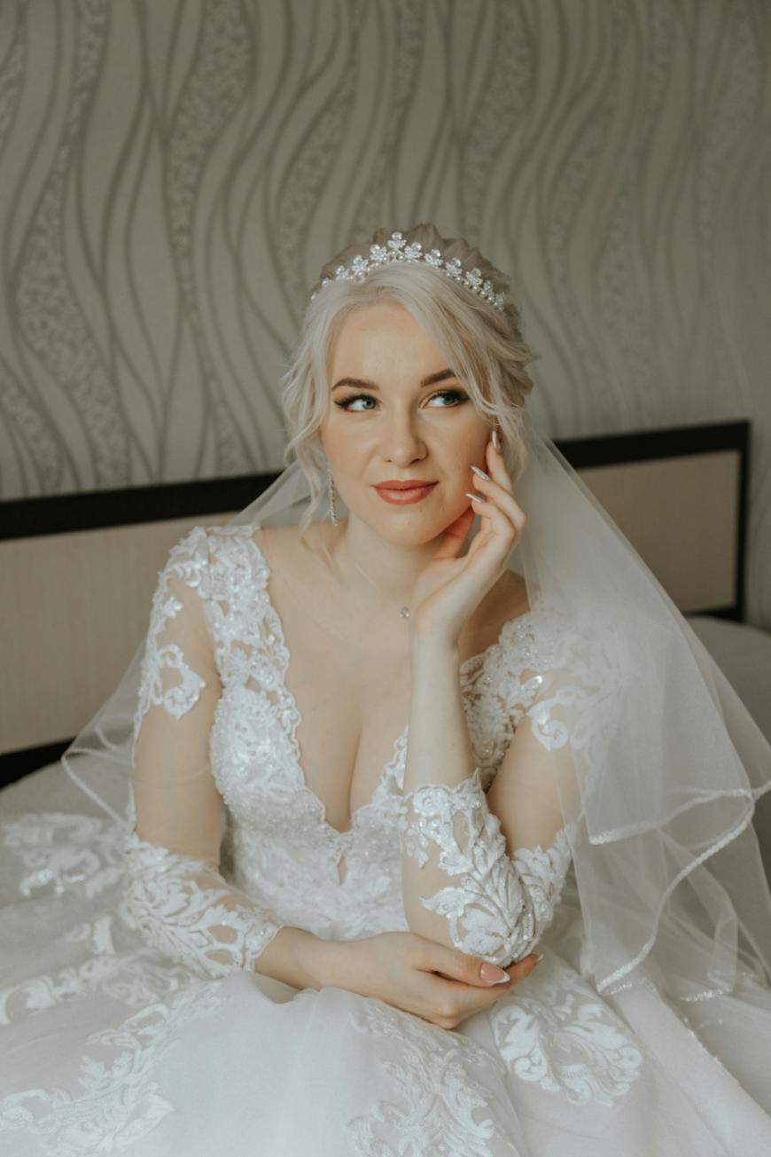 A bride with blonde hair, wearing a lace wedding dress, a jeweled headband, and a veil, sits thoughtfully with her hand resting on her face against a patterned background.