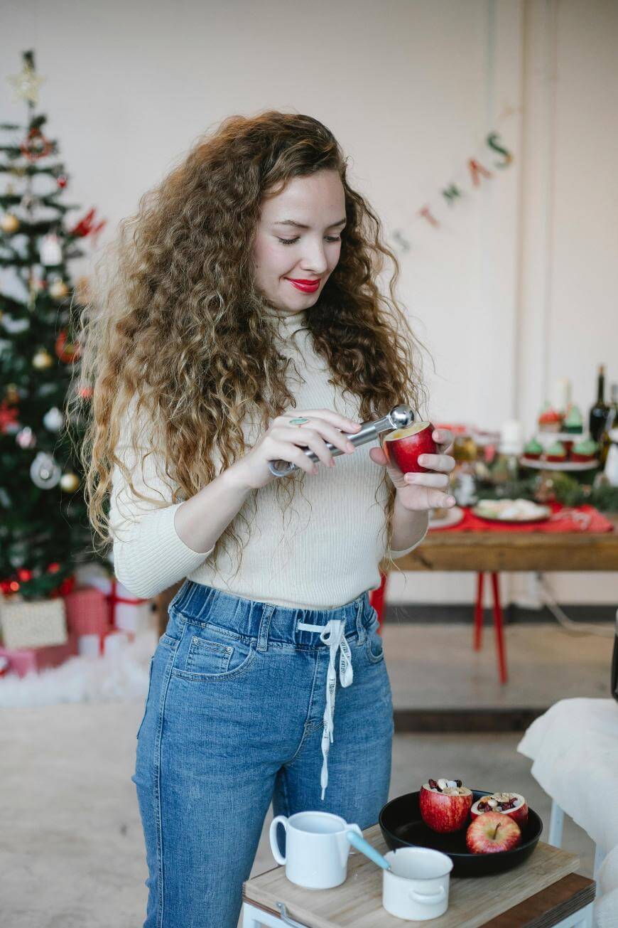 A woman with long curly hair cores a red apple in a festive room with a decorated Christmas tree and gifts in the background. A table with food and drinks is nearby. She wears a white sweater and blue jeans.