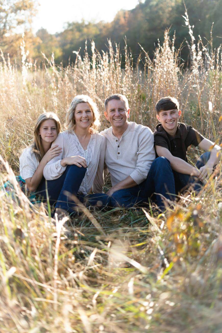 A family of four sitting together in a sunlit field of tall grass, smiling at the camera. The parents are in the center with their daughter and son seated on either side.