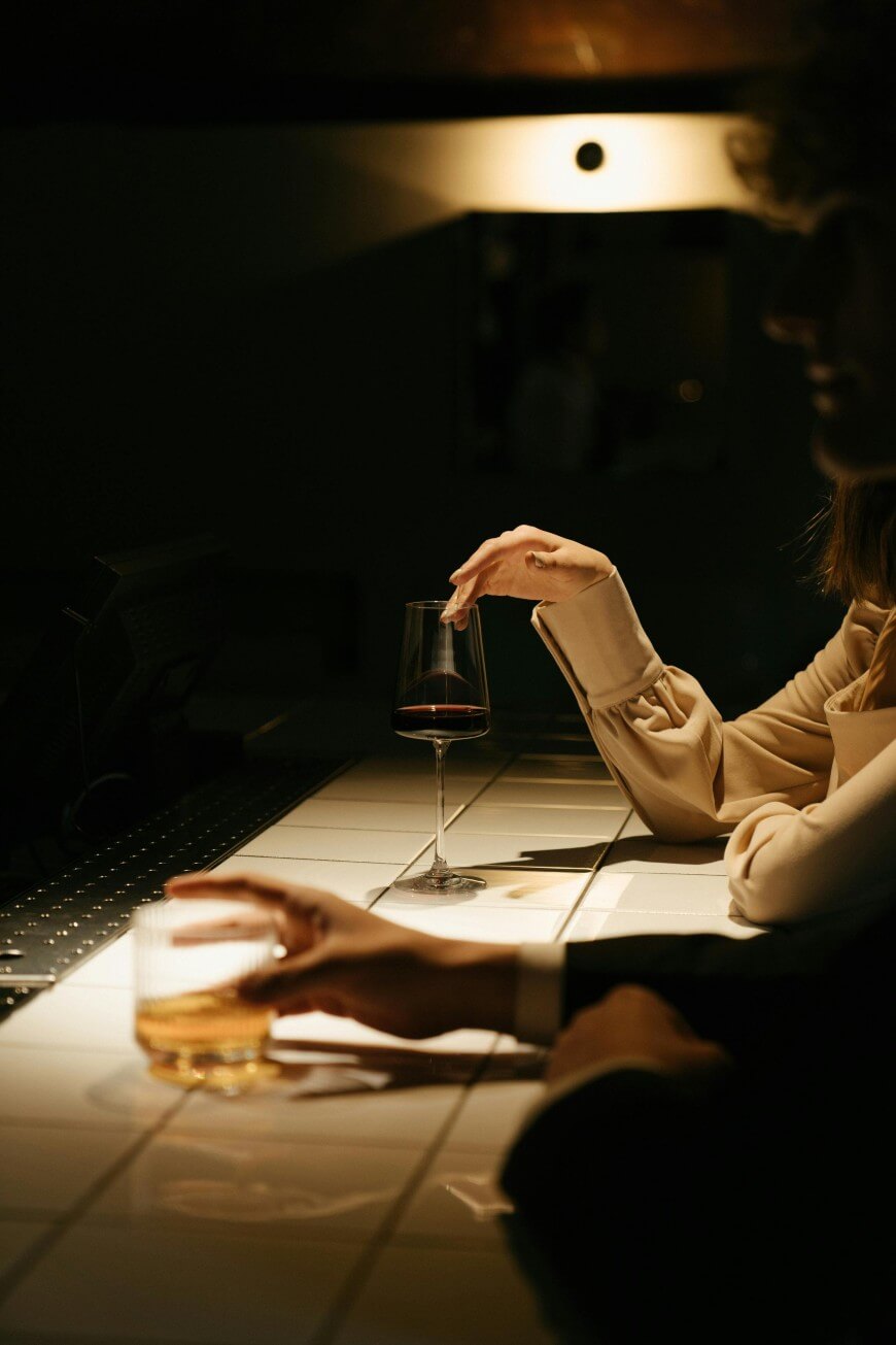 Two people sit at a dimly lit bar. One holds a glass of red wine with a cigarette, while the other rests their hand on a glass of amber-colored liquor. Only their hands and part of their arms are visible.