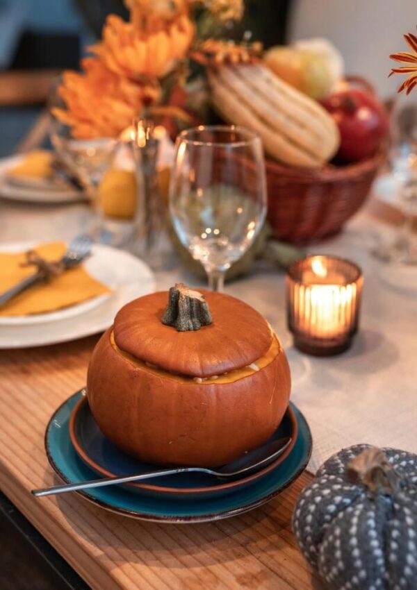 A small pumpkin filled with soup sits on a blue plate with a spoon beside it, on a festive autumn table set with plates, glasses, a candle, and a basket of gourds and apples in the background.
