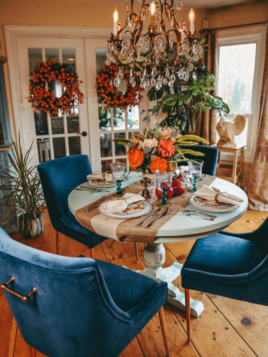 A cozy dining room with a round table set for four, blue velvet chairs, a floral centerpiece, and fall-themed decor, including orange wreaths on glass doors and a crystal chandelier above.