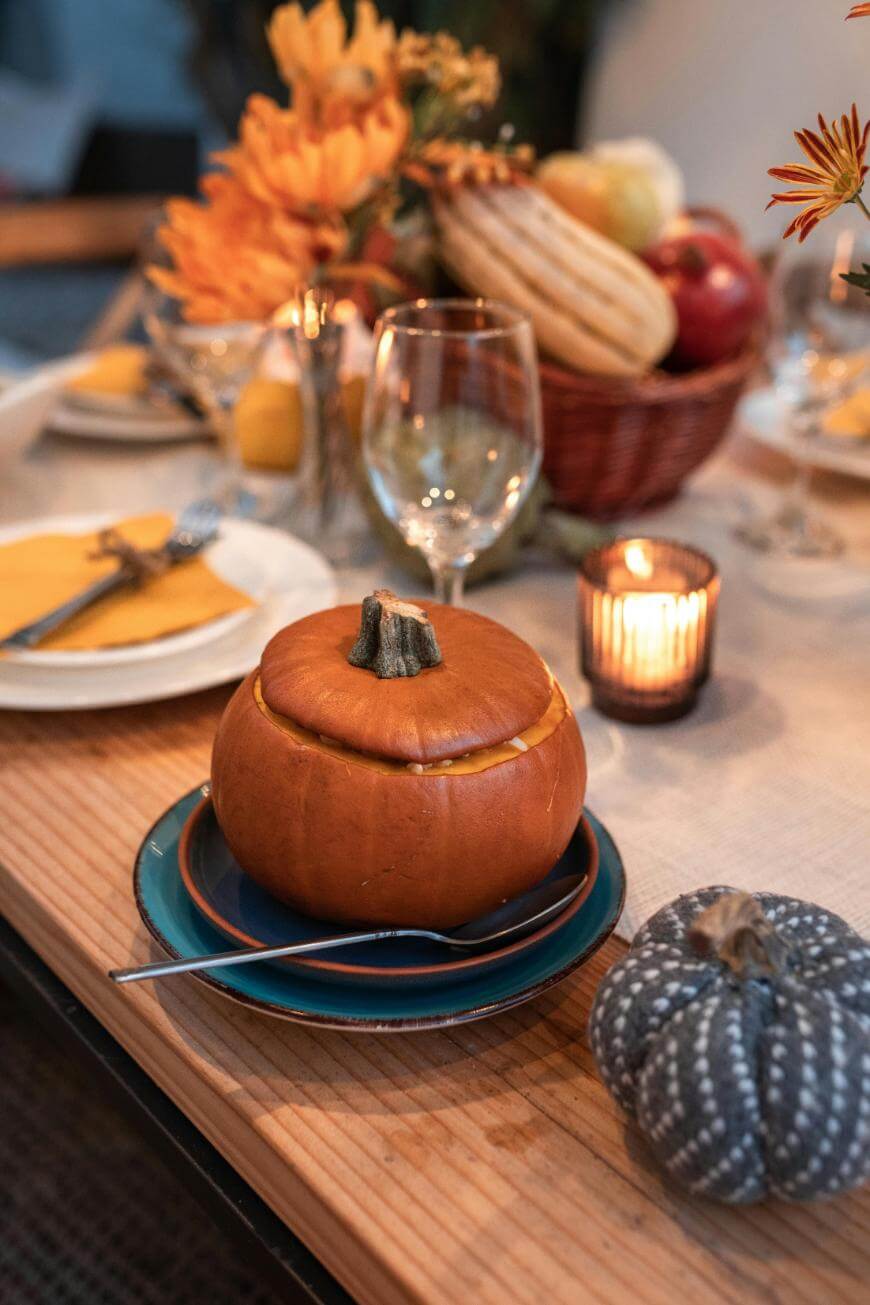 A festive table setting with a pumpkin soup served in a hollowed-out pumpkin, plates, cutlery, wine glasses, a candle, a basket of fruits, and autumn flowers as decoration.