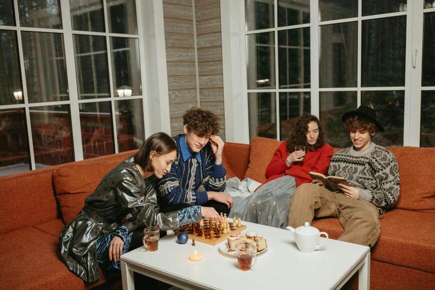 Four young adults sit on an orange sofa in a cozy room, with two playing chess and the other two relaxing, one reading a book and the other holding a mug. A white teapot and cups are on the coffee table in front of them.