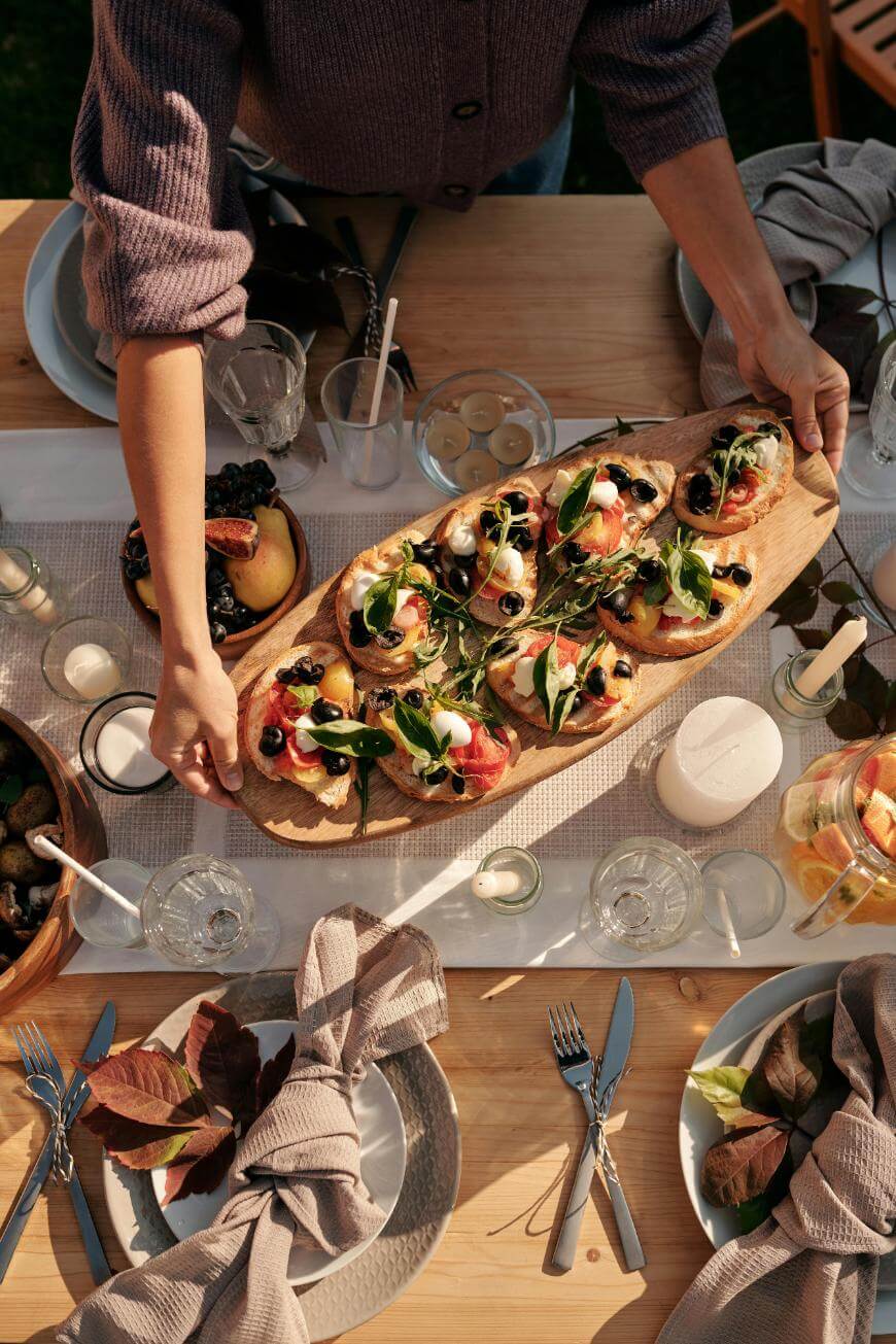 A person serves a wooden board with appetizers topped with greens and prosciutto at an outdoor table set with plates, glasses, candles, and autumn-themed decor.
