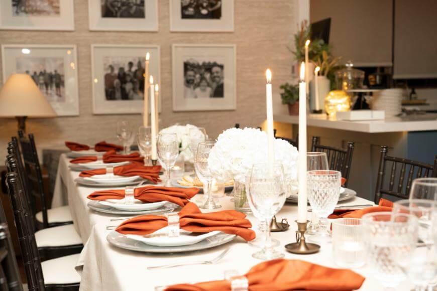 A dining table is set for a formal meal with white plates, crystal glasses, orange napkins, white floral centerpieces, and tall lit candles. Framed photos hang on the wall in the background.