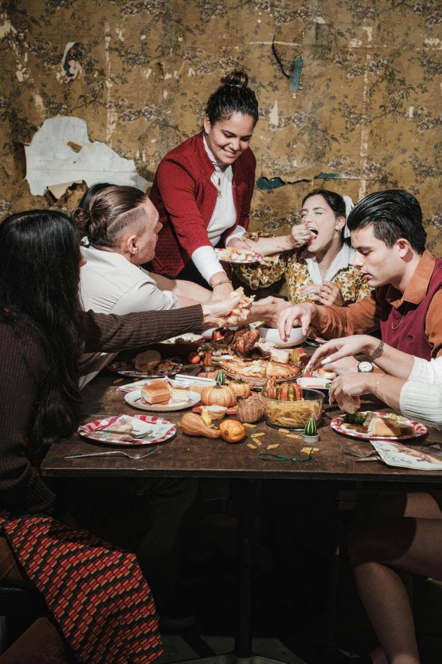 A group of five people sitting around a dining table sharing food and laughing, with various dishes and vegetables on the wooden table. One person is serving food while others enjoy the meal in a cozy setting.