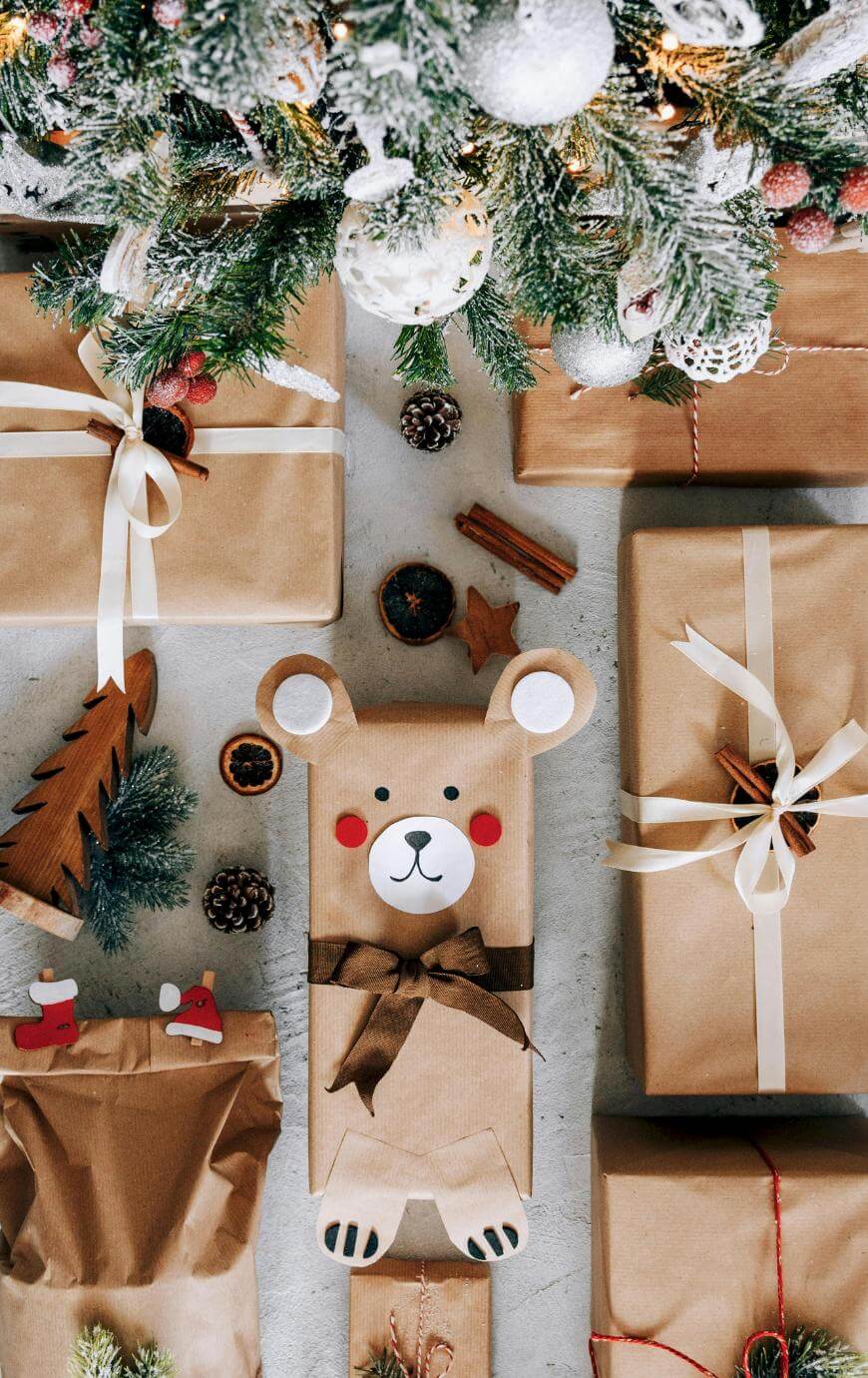 A group of brown-paper-wrapped Christmas presents with white ribbons are arranged under a decorated tree. One gift is wrapped to look like a bear, with a bow tie and a cute face. Pinecones and festive decorations are scattered around.